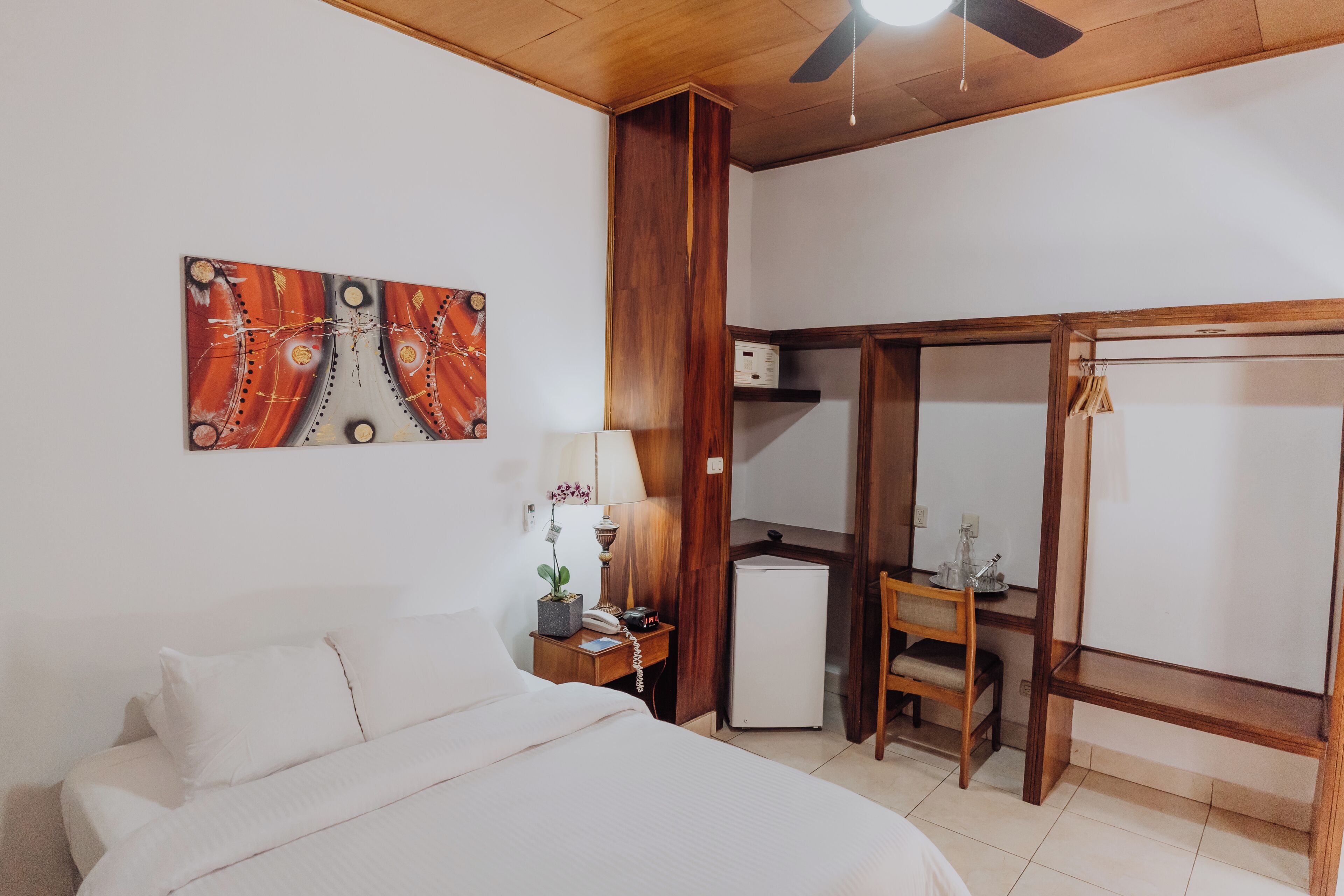 A bedroom with a bed, a desk, a chair, a small refrigerator, and a wardrobe.