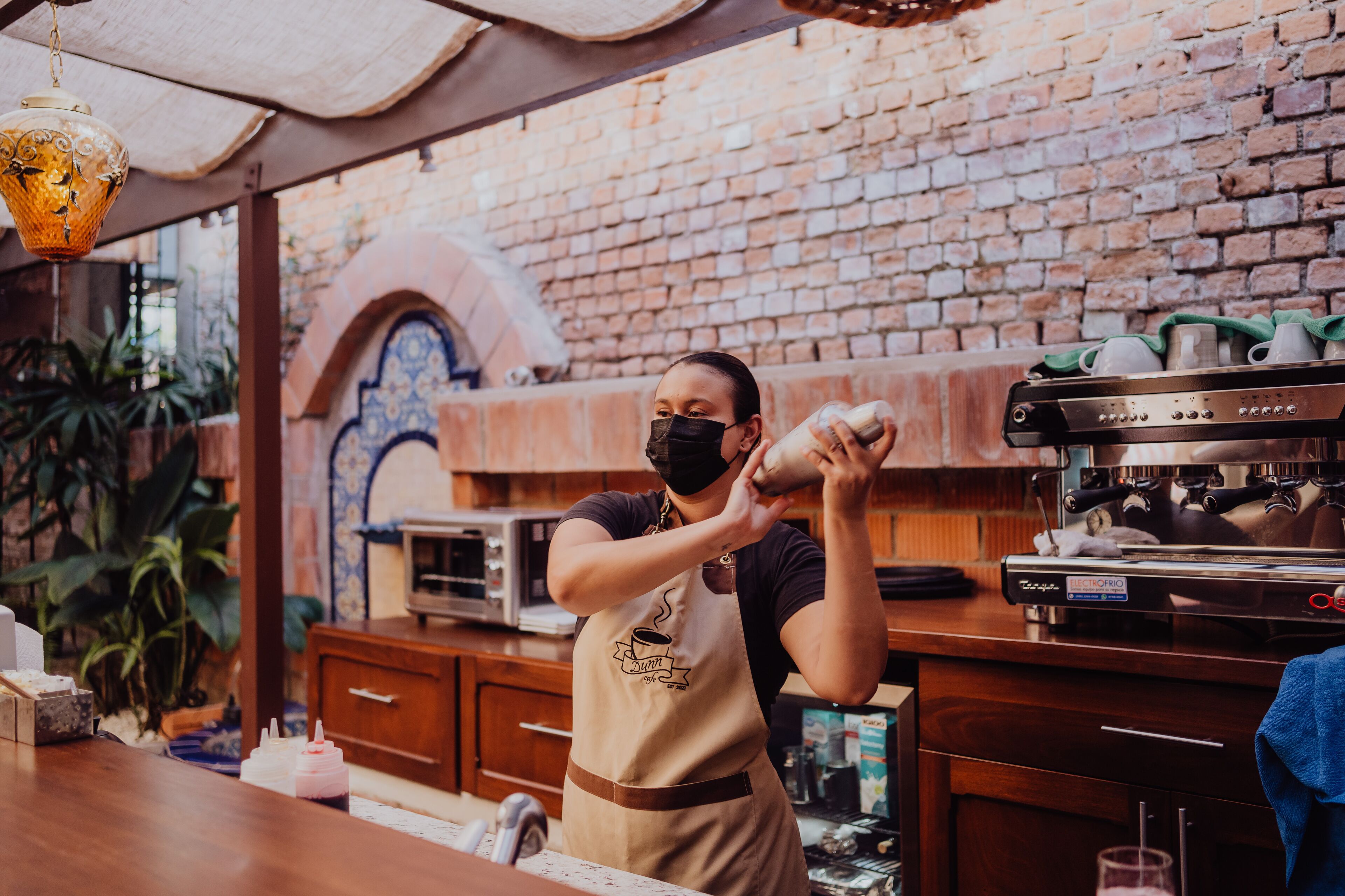 Una persona indossa un grembiule e prepara il caffè in una cucina con pareti in mattoni, mobili in legno e un lampadario.