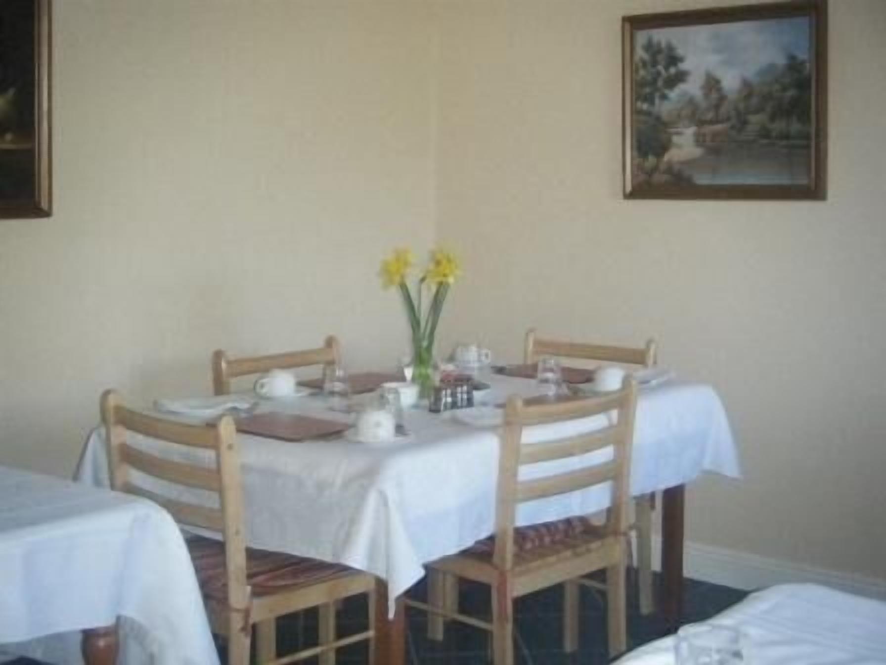 A dining area with a round table, four wooden chairs, and a white tablecloth. There is a vase with yellow flowers on the table.