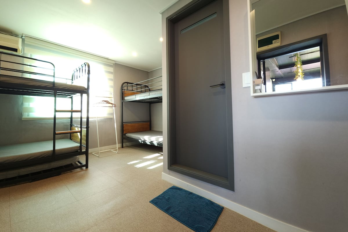 A room with bunk beds, a door, a window with blinds, and a small rug.