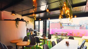 Um espaço de refeições interno com mesas e cadeiras, uma área de bar e vista para edifícios através de grandes janelas.