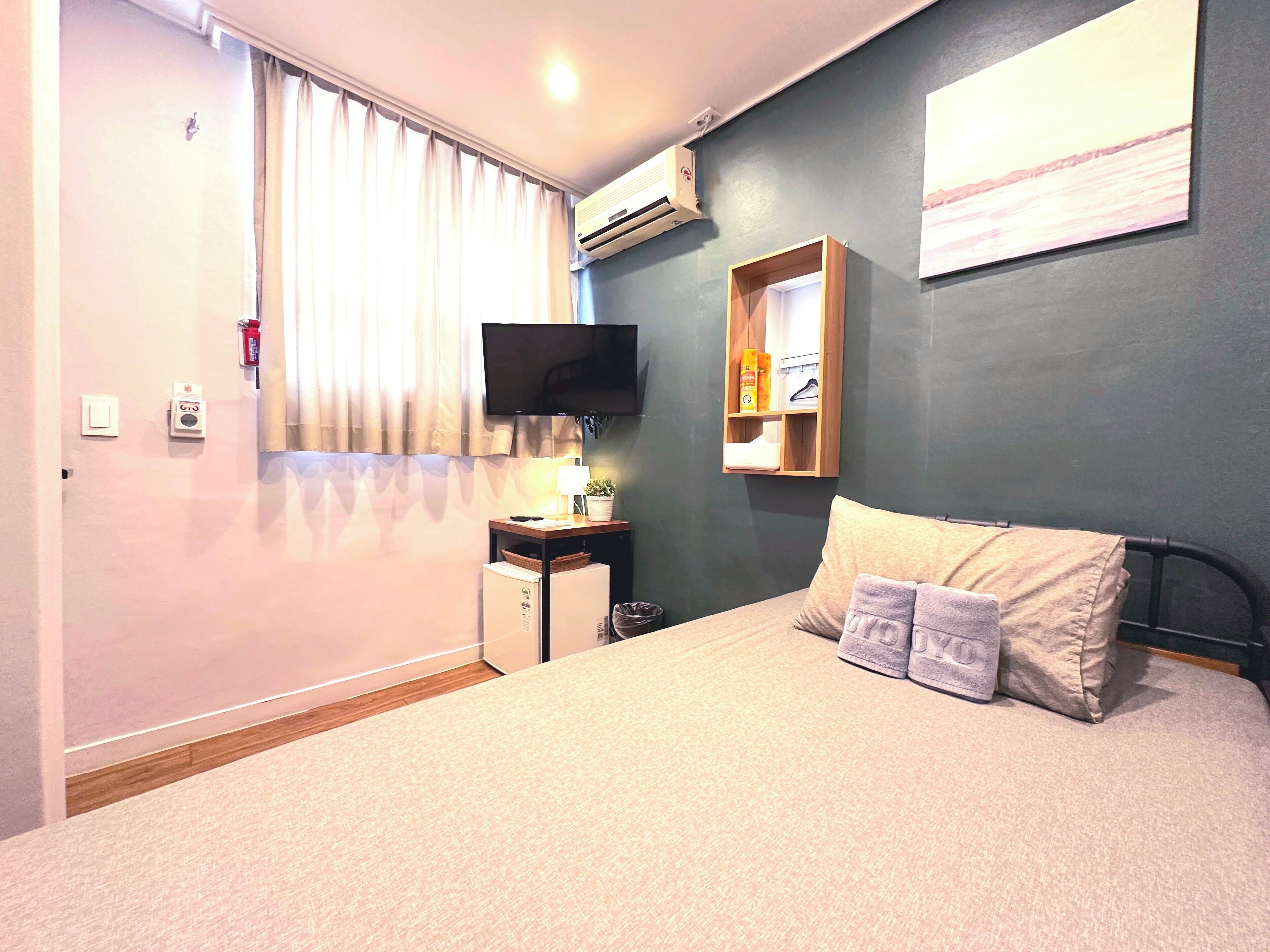 A bedroom with a bed, a TV, a small table with a lamp, and a shelf with toiletries.