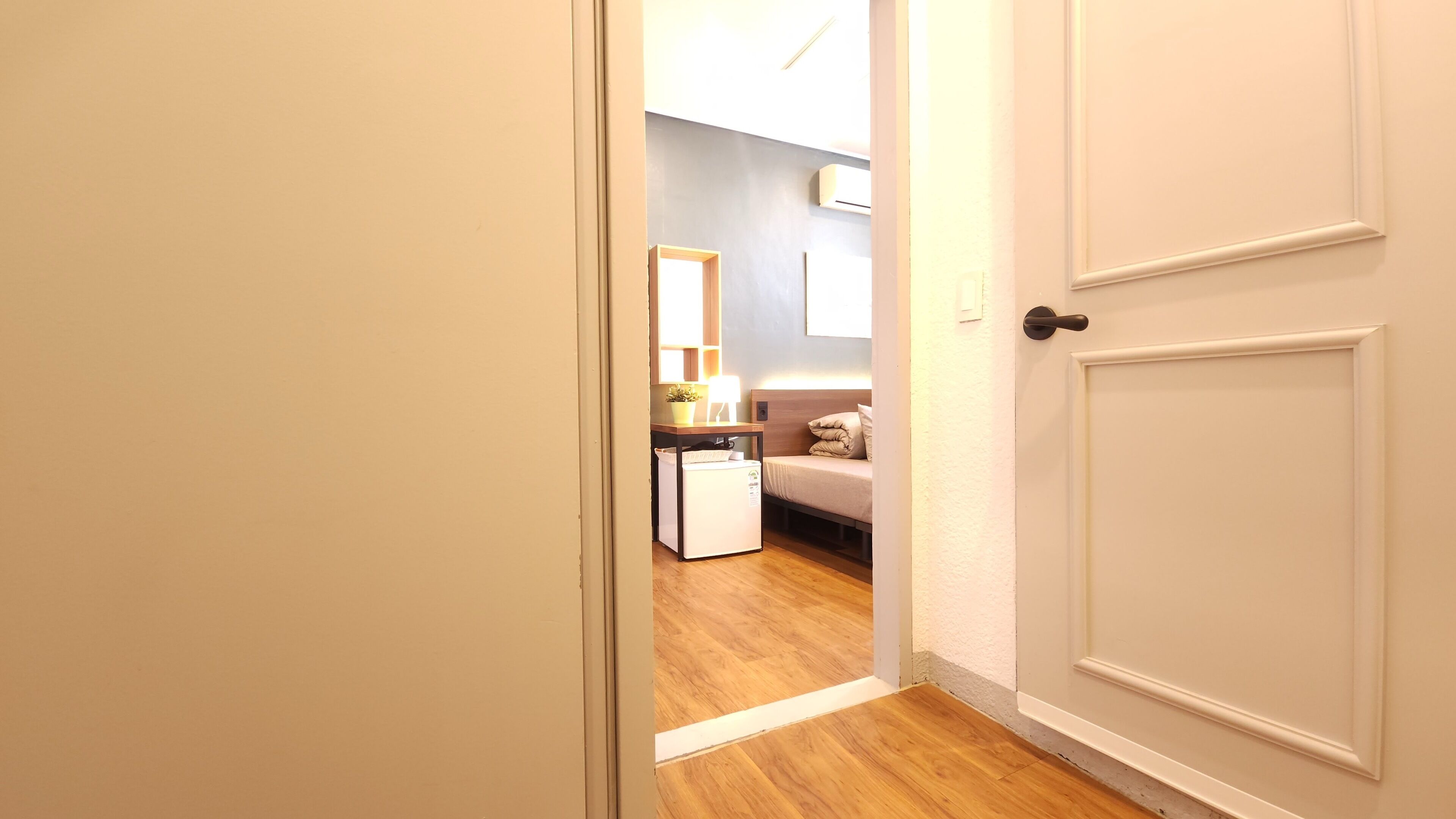 Un couloir étroit mène à une chambre où se trouvent un lit, une petite table et un climatiseur blanc fixé au mur.