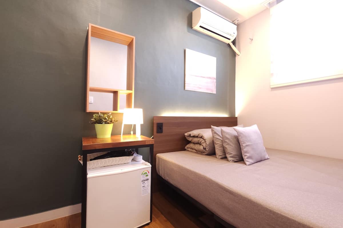 A compact bedroom with a bed, a small desk with a lamp, a potted plant, and a mini-fridge.