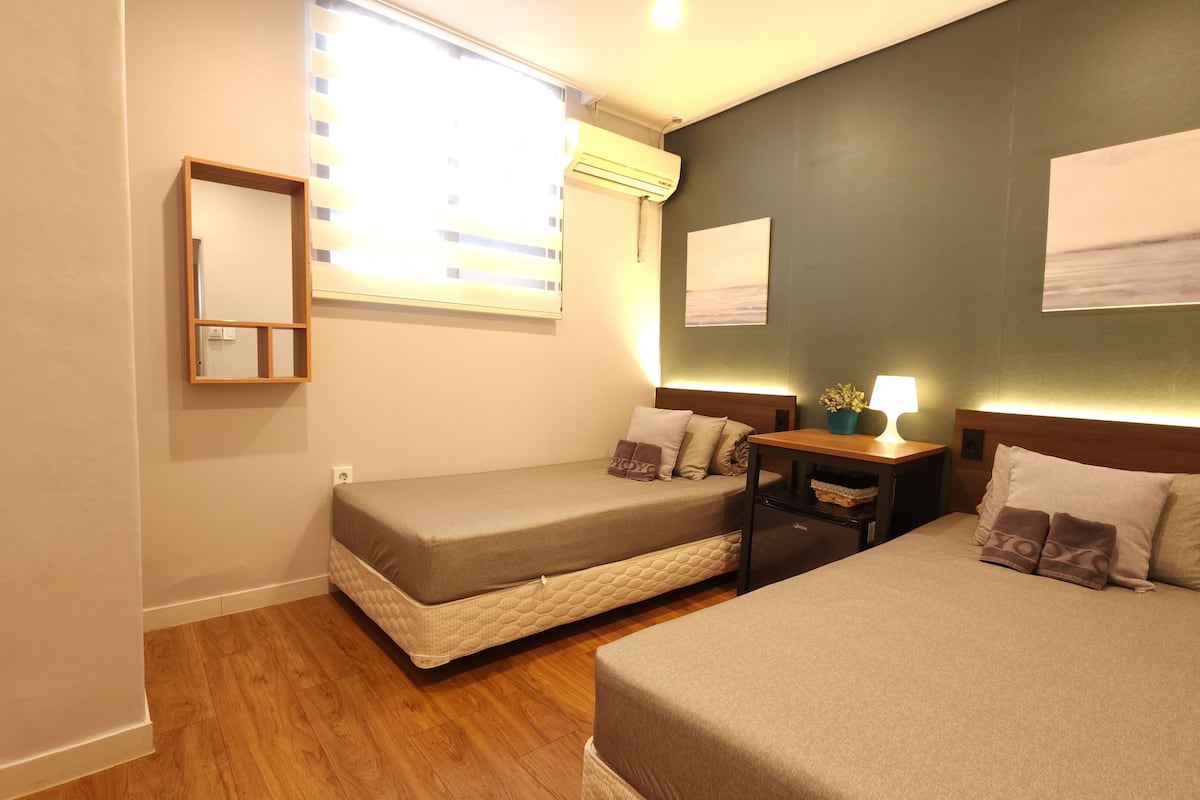 A hotel room with two beds, a bedside table with a lamp, a mirror, and a window with blinds.