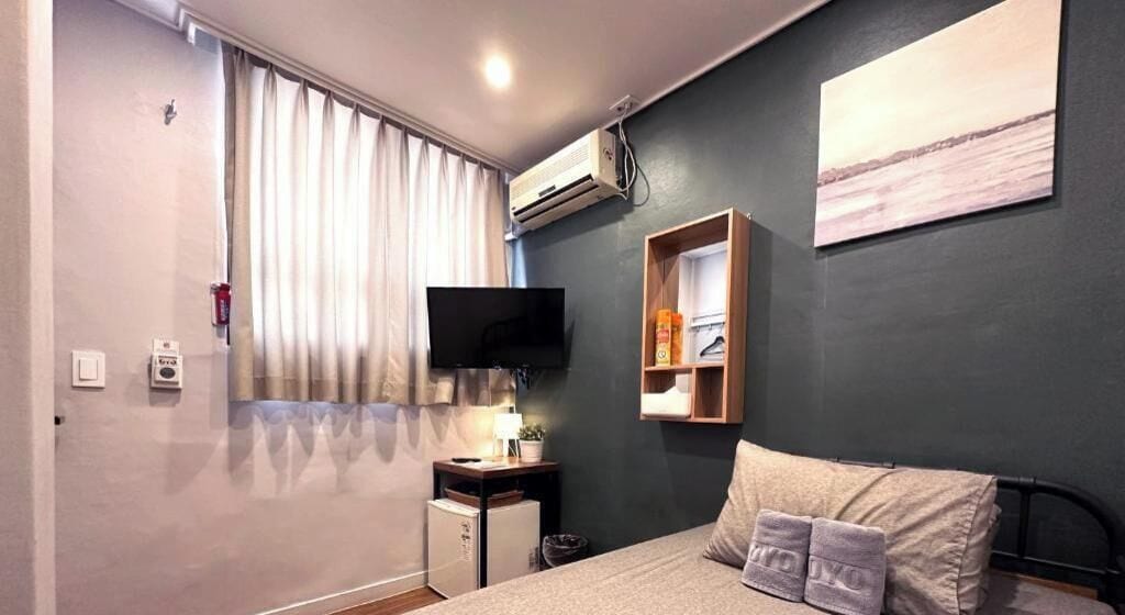 A bedroom with a bed, a TV, a small desk, a shelf, and a window with curtains.