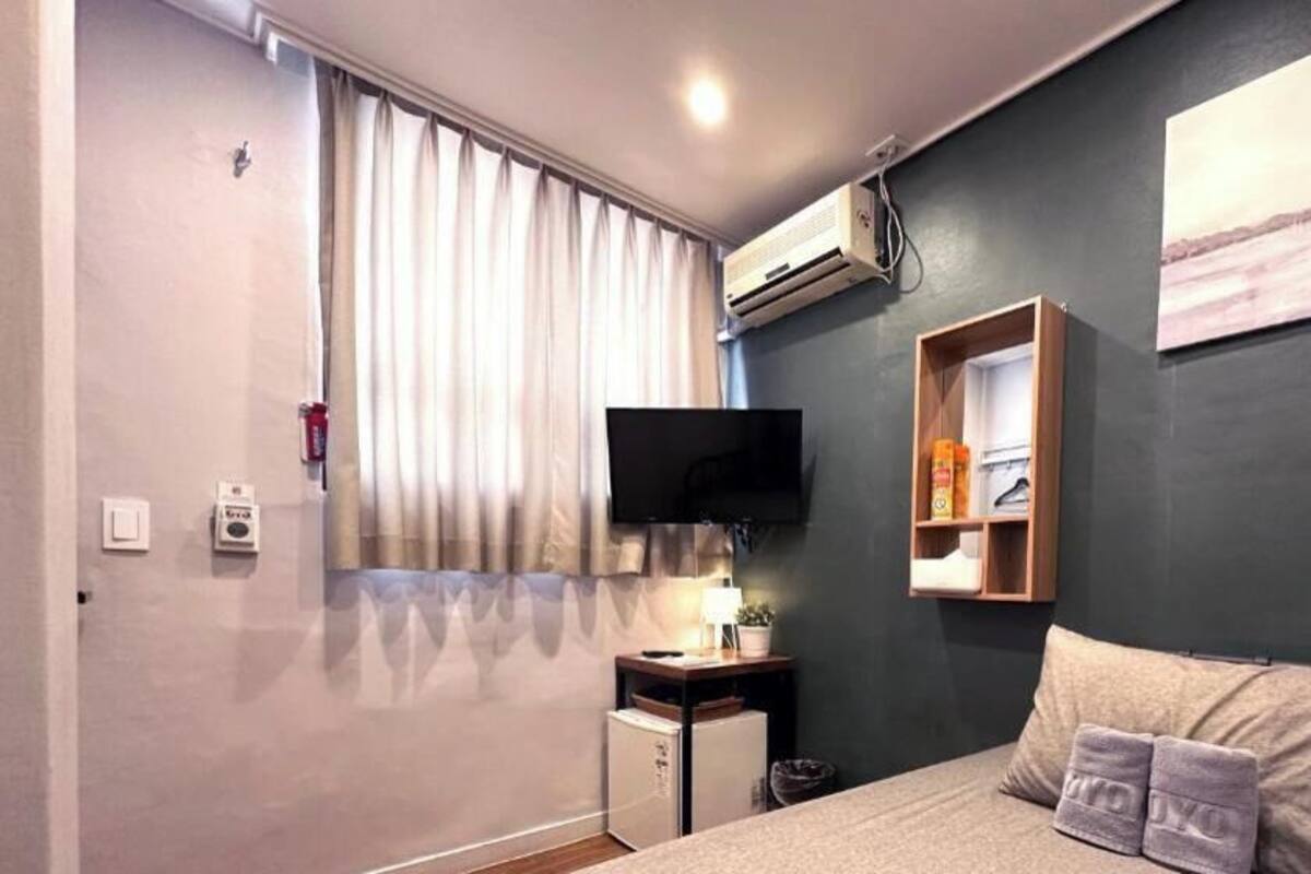 A bedroom with a bed, a TV, a small desk, a shelf, and a window with curtains.