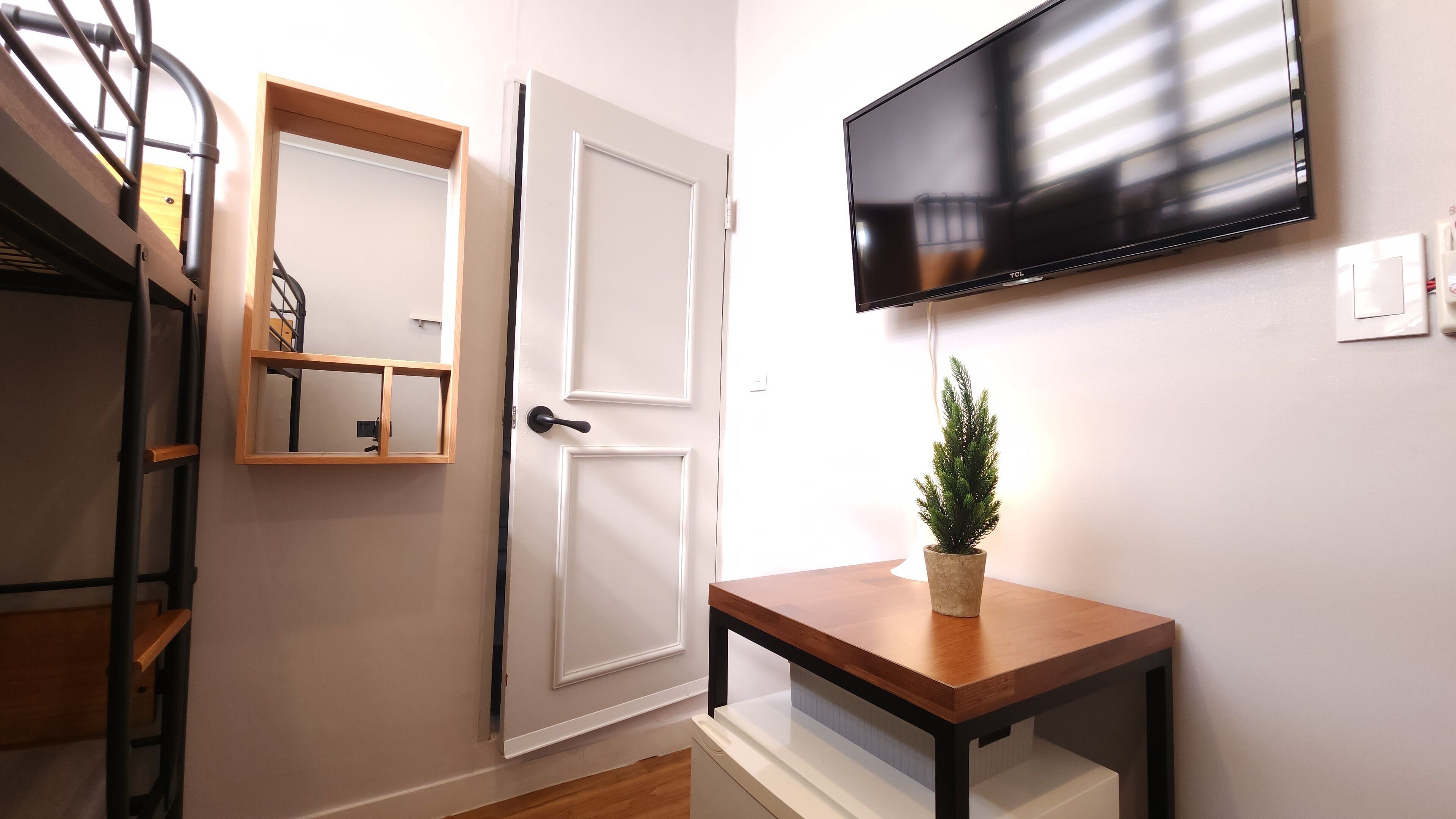 Una habitación con un escritorio de madera, una planta en maceta, un televisor montado y una puerta con espejo.