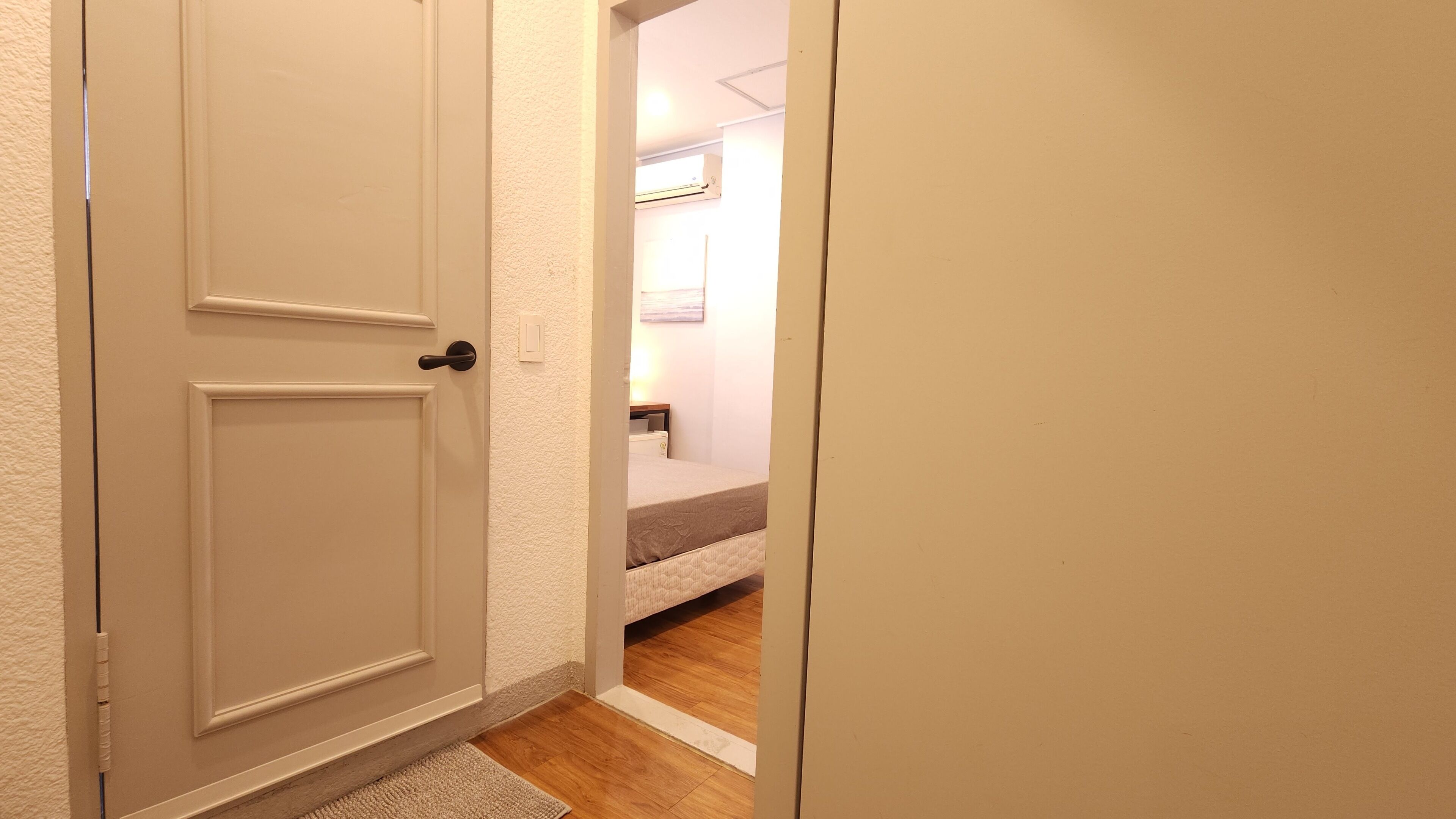 Un pasillo estrecho que lleva a un dormitorio con una cama, una mesita de noche y un aire acondicionado instalado en la pared.