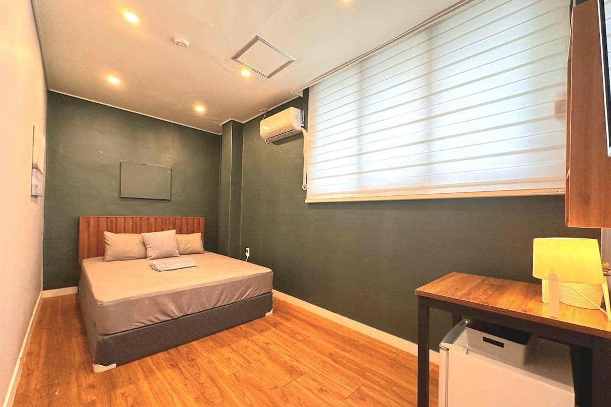 A bedroom with a bed, a desk, a lamp, and a window with blinds.