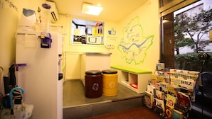 Une petite pièce équipée d’un réfrigérateur, d’un four à micro-ondes et d’une machine à café. On y trouve deux poubelles et une petite étagère avec des livres. Une carte est accrochée au mur, et une fenêtre donne sur un arbre à l’extérieur.
