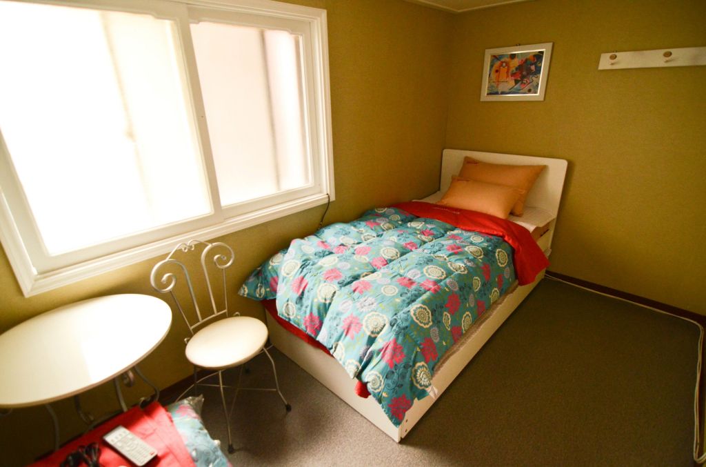 A small, well-lit bedroom with a single bed, a small table, and a chair.