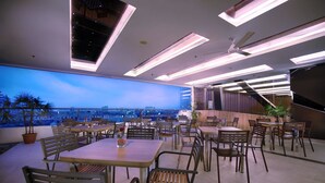 Een modern restaurant met meerdere tafels en stoelen, uitzicht op de stad en een plafond met lineaire verlichtingsarmaturen.
