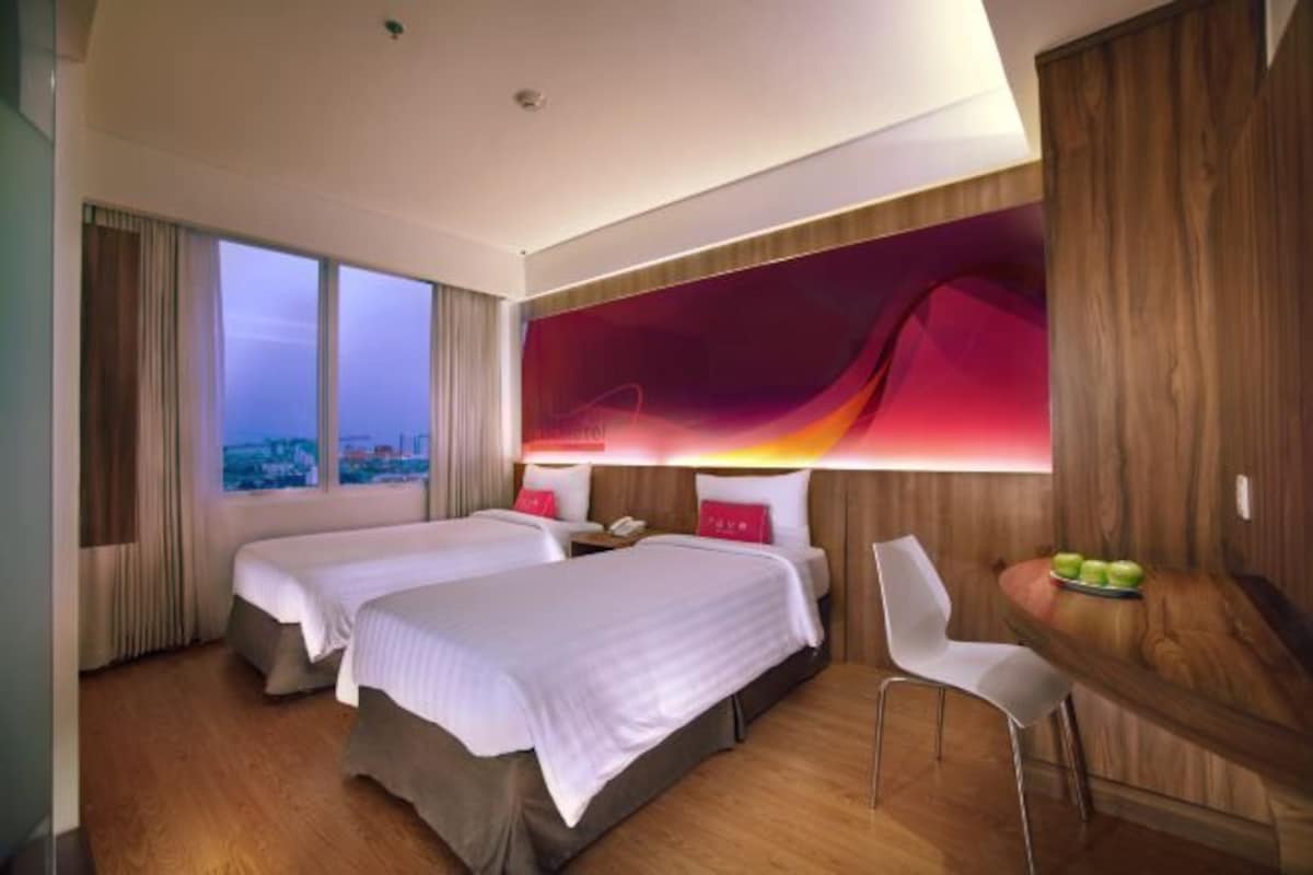 Une chambre d’hôtel avec deux lits, une grande fenêtre donnant sur la ville, une œuvre d’art moderne accrochée au mur et une petite table avec des fruits.