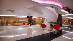 Een moderne hotellobby met een receptiebalie, rode stoelen en een groot digitaal bord waarop de naam van het hotel staat.