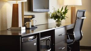Habitación de hotel con televisor de pantalla plana montado en la pared, escritorio con cafetera, una silla y un florero con flores.