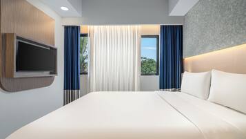 A modern hotel room with a large bed, a flat-screen TV mounted on the wall, and a view of greenery outside.