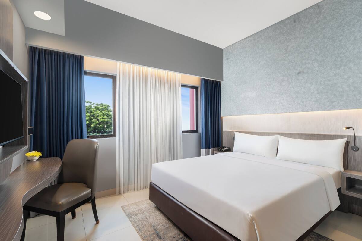 Een moderne hotelkamer met een groot bed, een bureau met stoel, een televisie en uitzicht op groen.