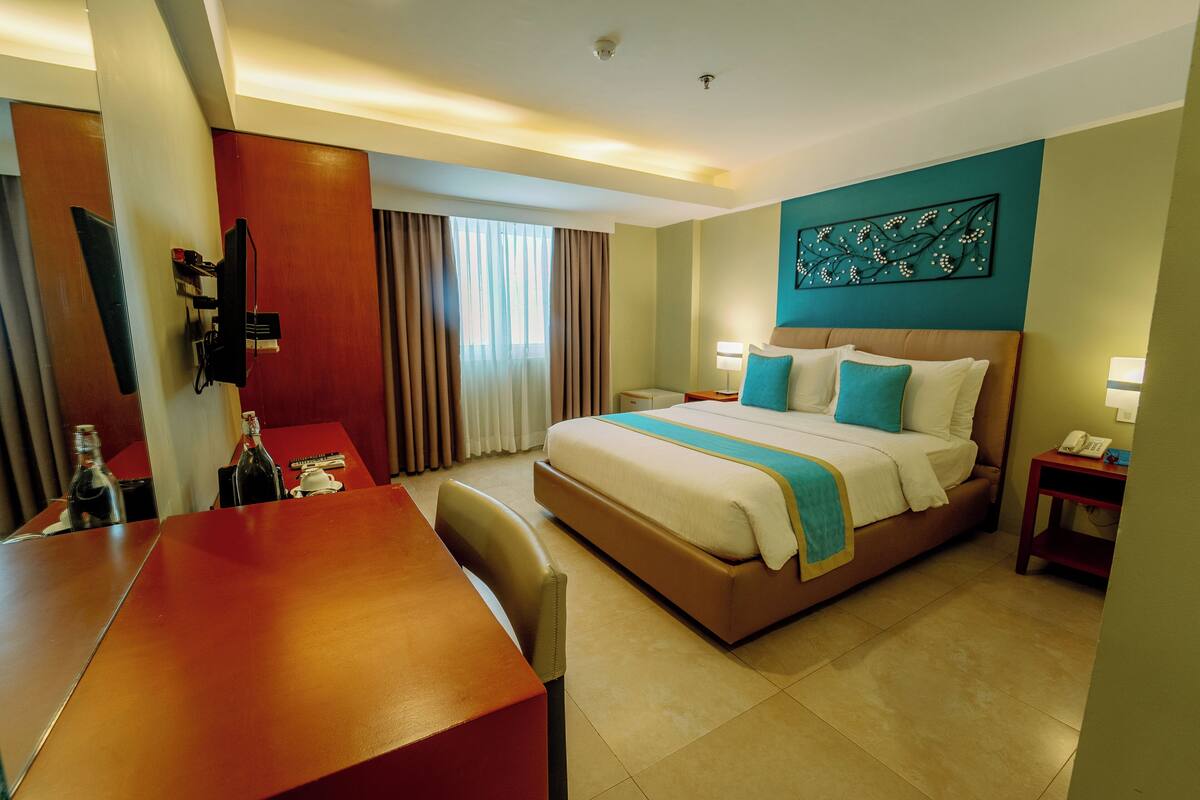 Habitación de hotel con una cama grande, un escritorio y una silla. Cuenta con una ventana con cortinas, un televisor montado en la pared y un cuadro sobre la cama.