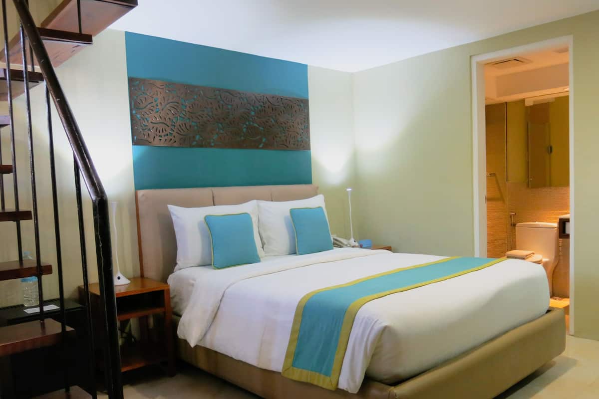 Habitación de hotel con cama, mesita de noche, lámpara y baño visible a través de una puerta abierta.