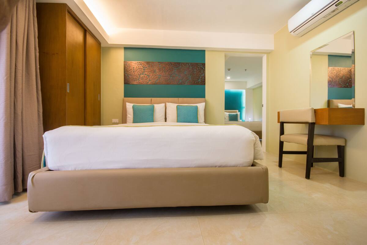 Una habitación de hotel moderna con una cama grande, un espacio de descanso con una silla, un espejo y un baño visible a través de una puerta abierta.