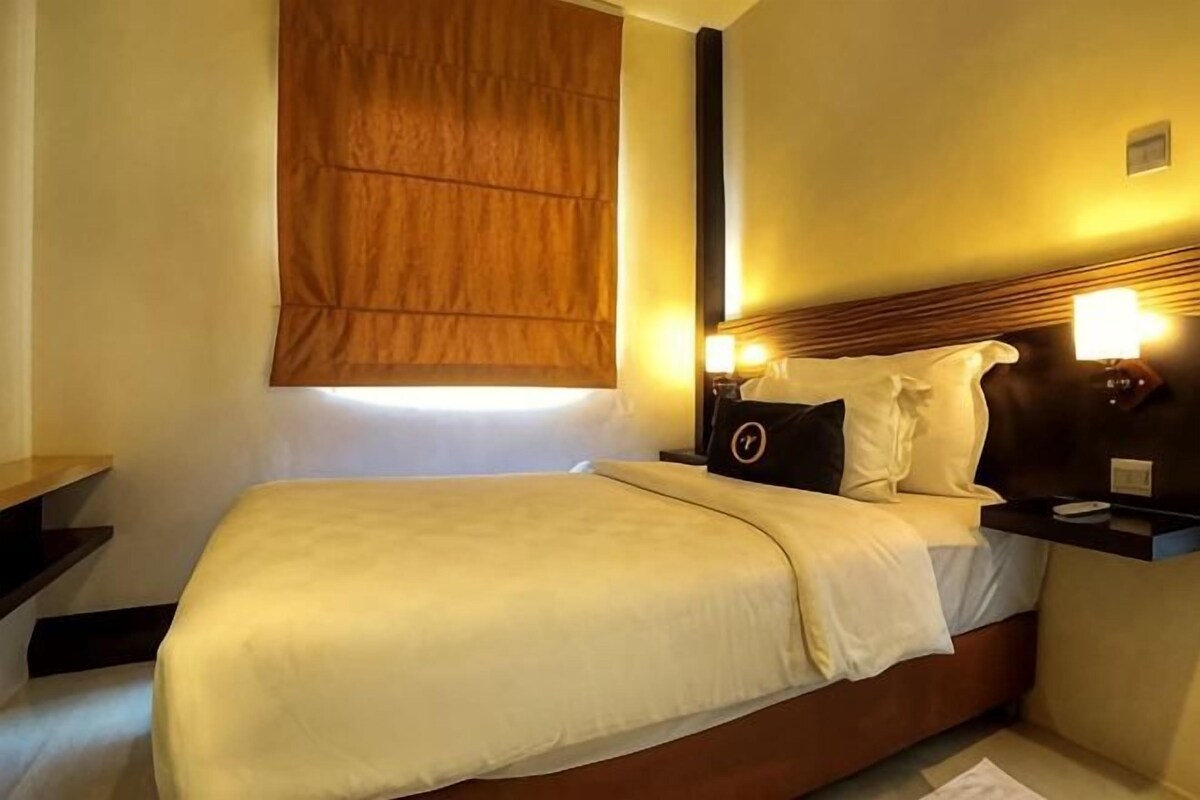 A hotel room with a bed, bedside tables, a nightstand, a window with a blind, and a wall-mounted lamp.