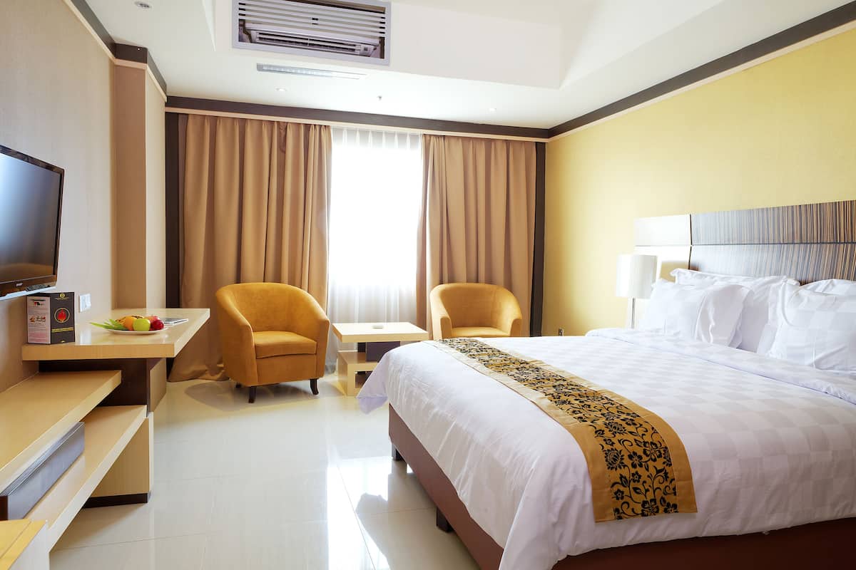 Une chambre d’hôtel comprenant un lit, deux fauteuils, un bureau avec un ordinateur portable, un téléviseur à écran plat et un panier de fruits.
