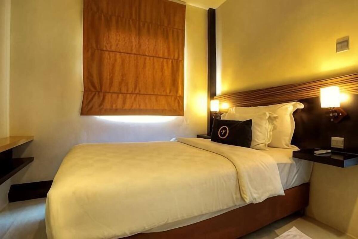 Une chambre d’hôtel comprenant un lit, des tables de chevet, une petite table, une fenêtre avec un store et une lampe fixée au mur.