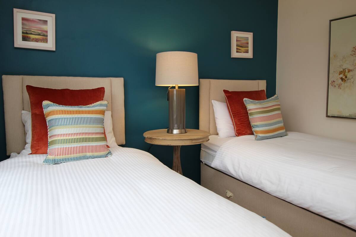Een hotelkamer met twee bedden, elk met een hoofdeinde, een nachtkastje met een lamp, en kunstwerken aan de muur.