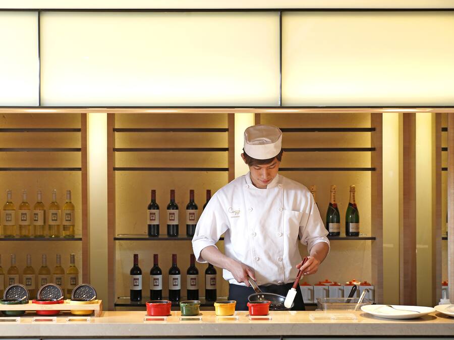 A chef preparing food in a well-lit kitchen with wine bottles in the background.