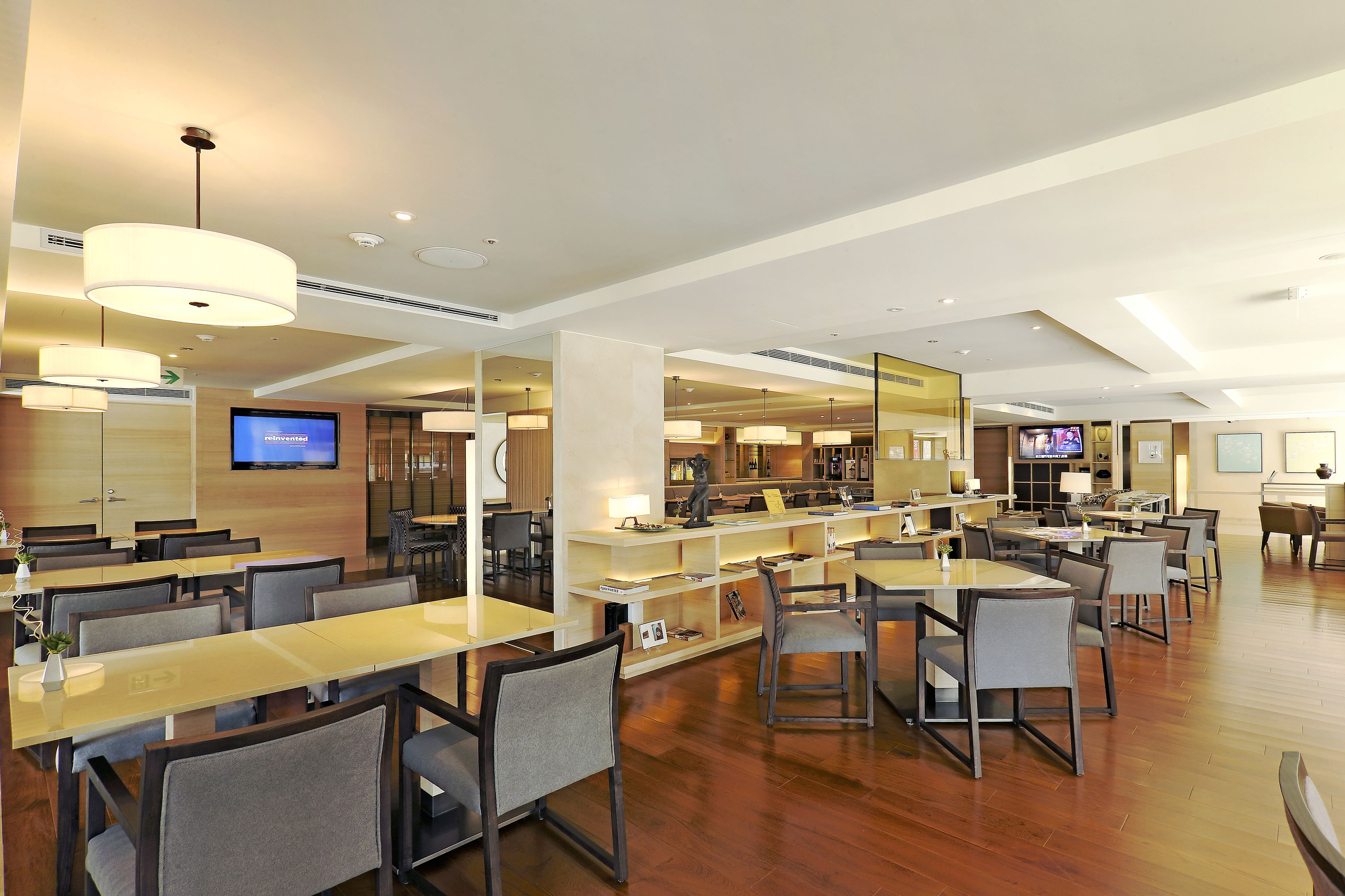 A modern hotel lobby with a long reception desk, multiple seating areas, and a television screen displaying 'reinvented'.