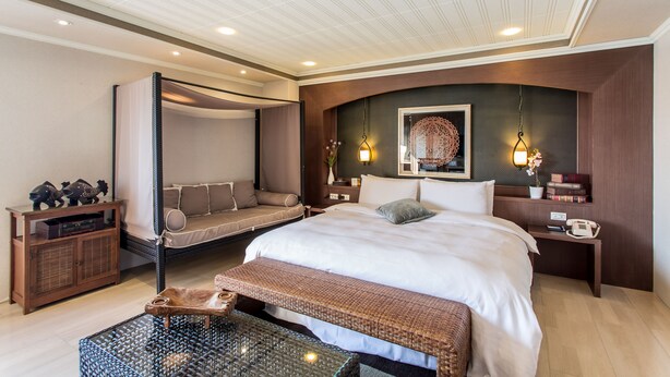 Una habitación de hotel moderna con una cama grande, un sillón cama, una mesita pequeña y un banco.