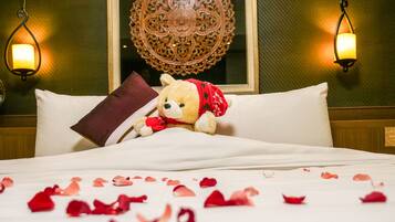 A bed with rose petals scattered on it, a teddy bear wearing a red scarf, and two wall-mounted lamps.