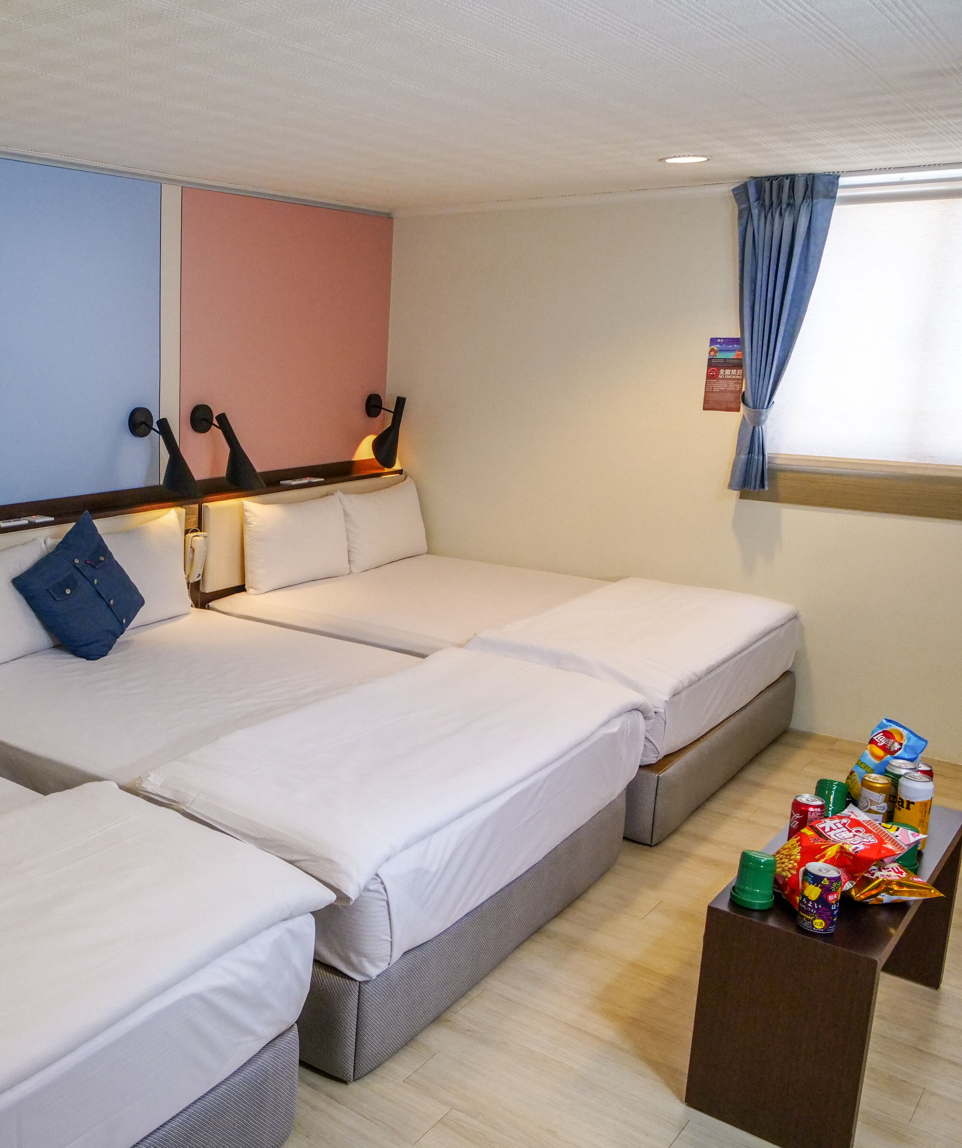 A hotel room with three beds, a small table with snacks, and a window with curtains.