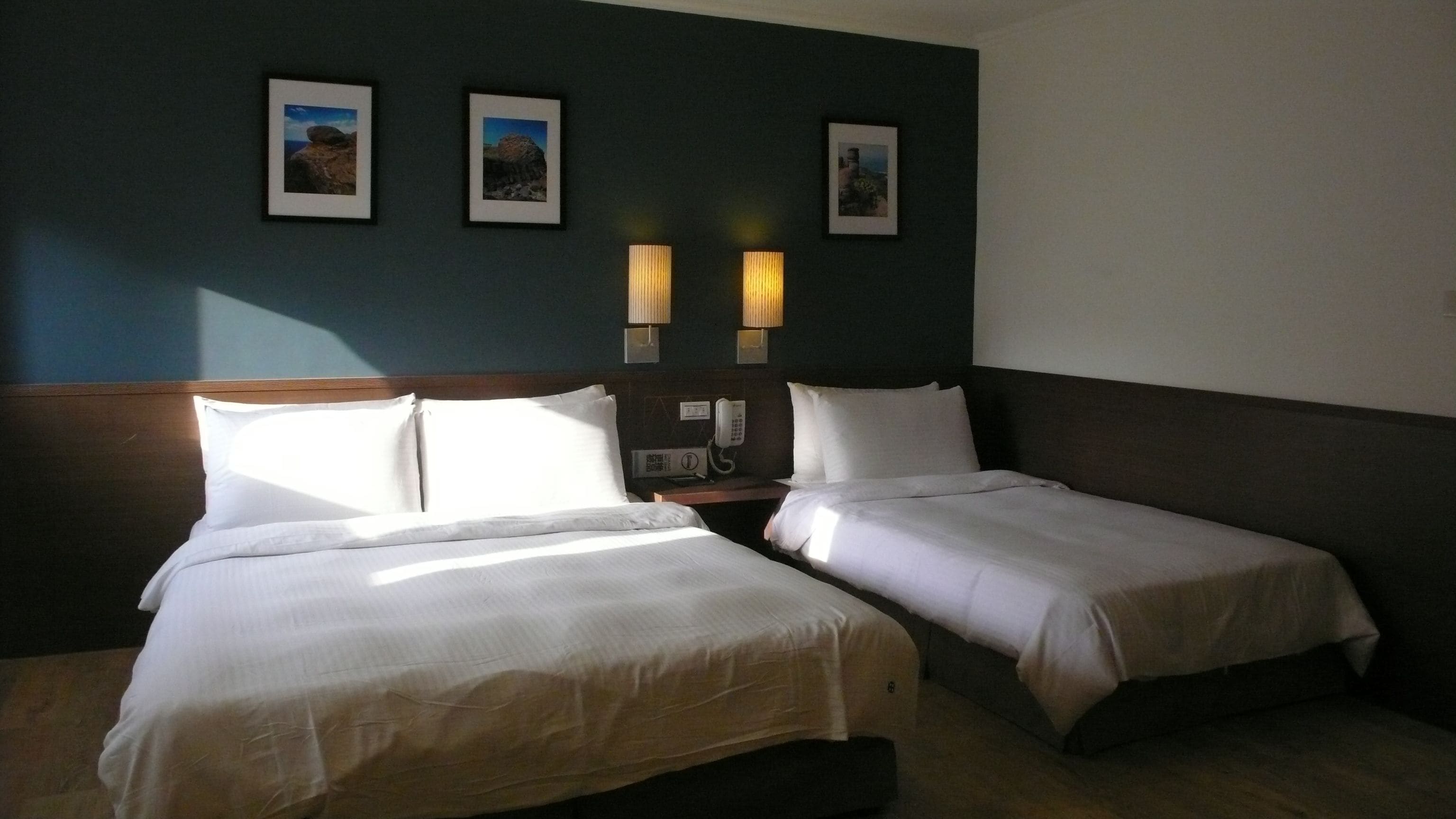 Habitación de hotel con dos camas, cabecero de madera, mesita de noche con teléfono y tres cuadros enmarcados en la pared.
