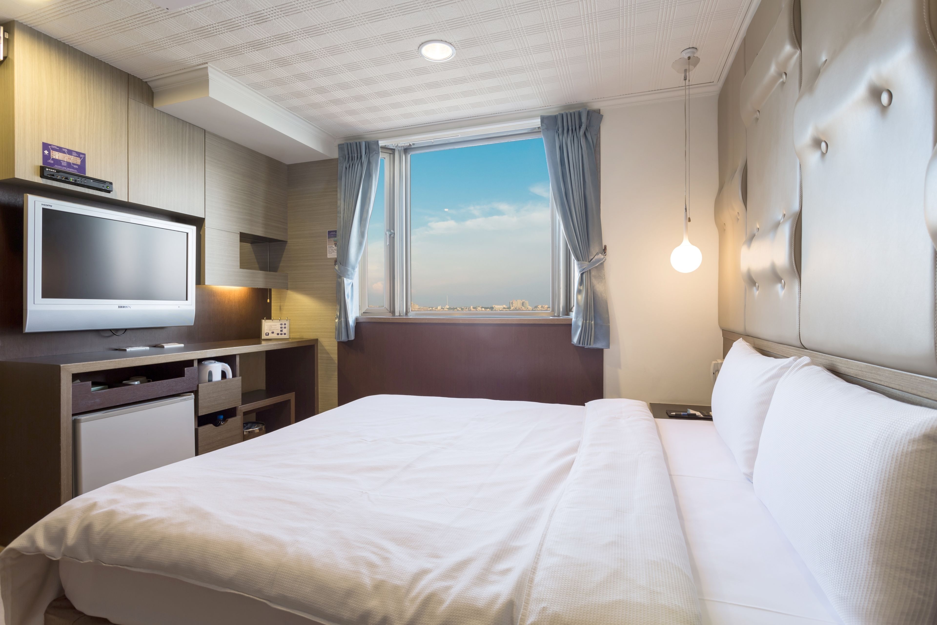 Quarto de hotel com cama, televisão, secretária com computador e janela com cortinas.
