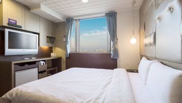 Quarto de hotel com cama, televisão, secretária com computador e janela com cortinas.