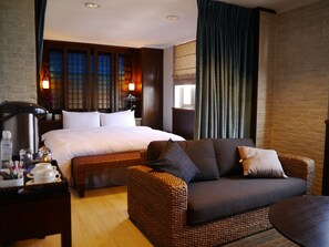 Habitación de hotel con una cama grande, un sofá de mimbre, una mesita de noche con artículos de aseo y una ventana con cortinas.