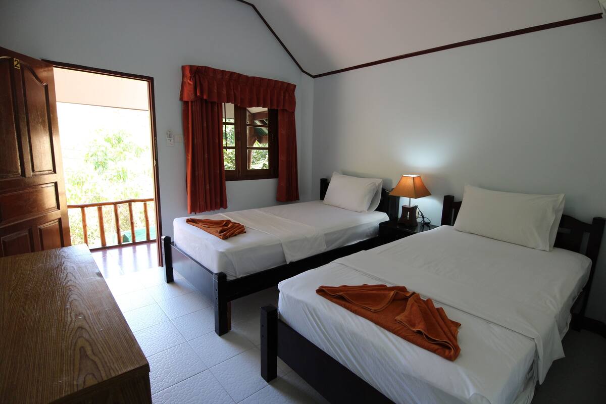 A room with two single beds, a wooden desk, a window with curtains, and a nightstand with a lamp.