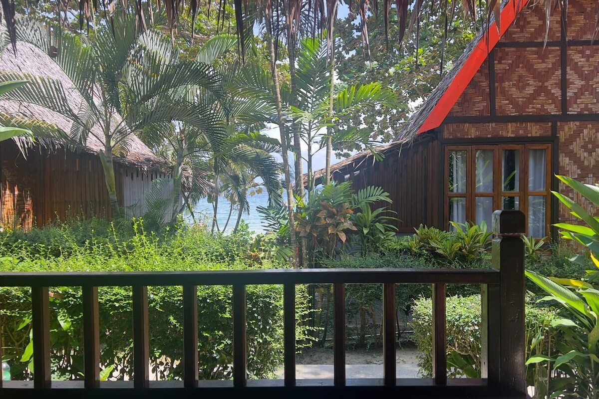 Una casa de madera tradicional con techo de paja, rodeada de exuberante vegetación tropical y vista al mar.