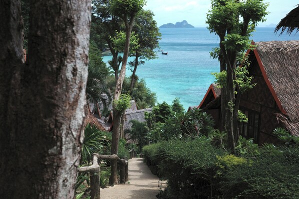 A pathway leading to a thatched-roof building surrounded by lush greenery and overlooking a calm sea with boats.