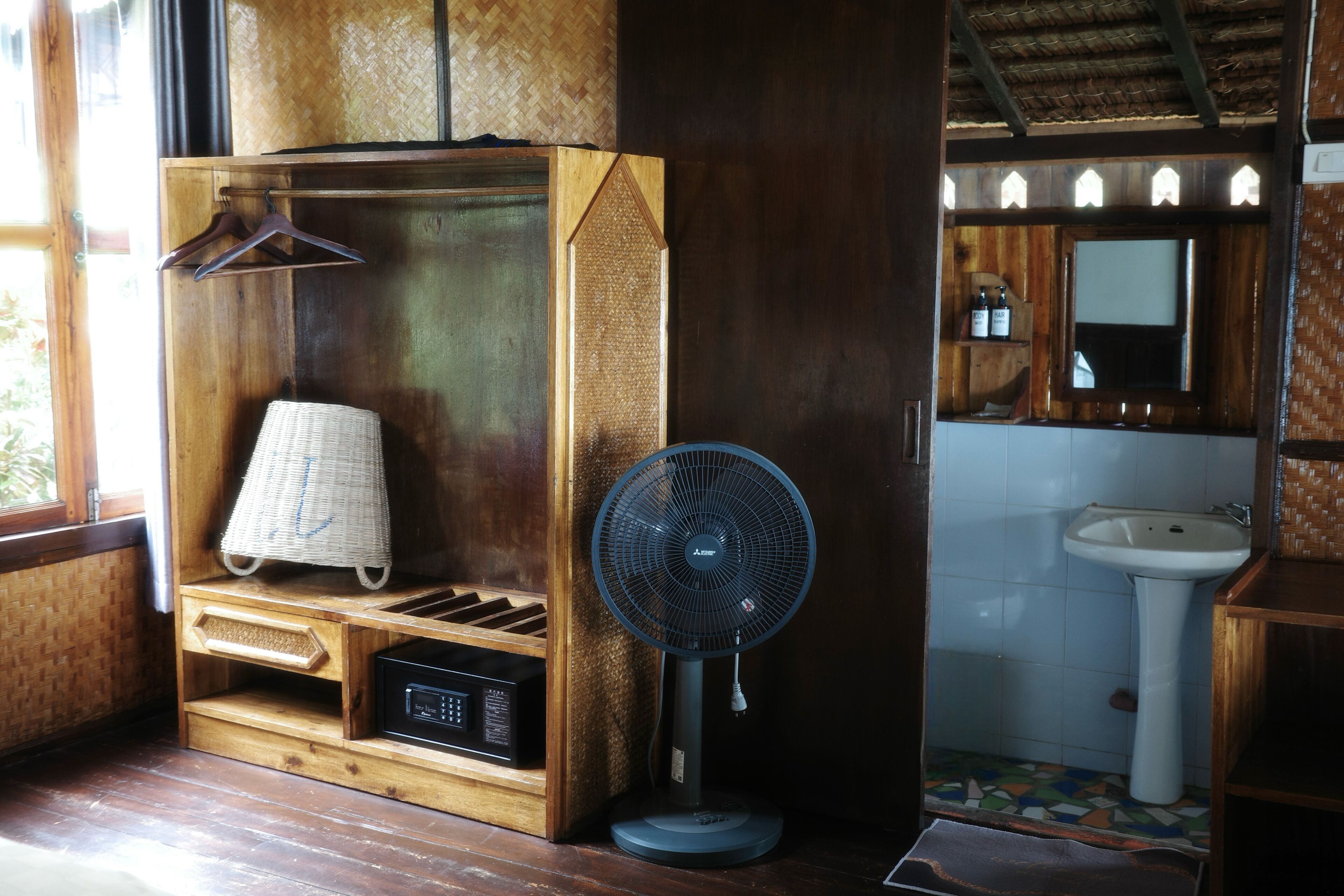 A wooden cabinet with a radio, a hanging laundry basket, and a standing fan in a bathroom.