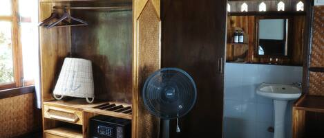 A wooden cabinet with a radio, a hanging laundry basket, and a standing fan in a bathroom.