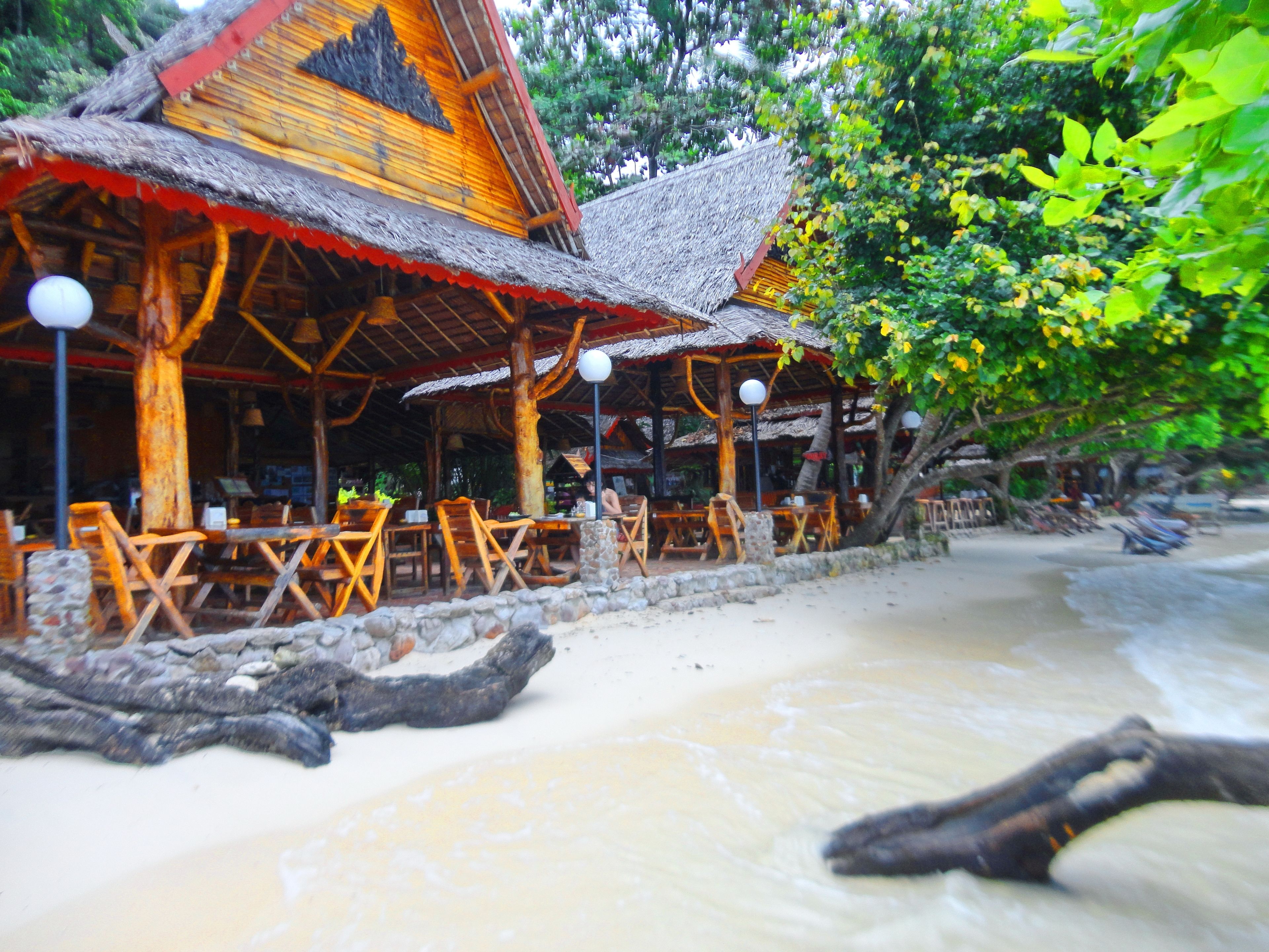 A beachside restaurant with wooden tables and chairs, a thatched roof, and stone walls.