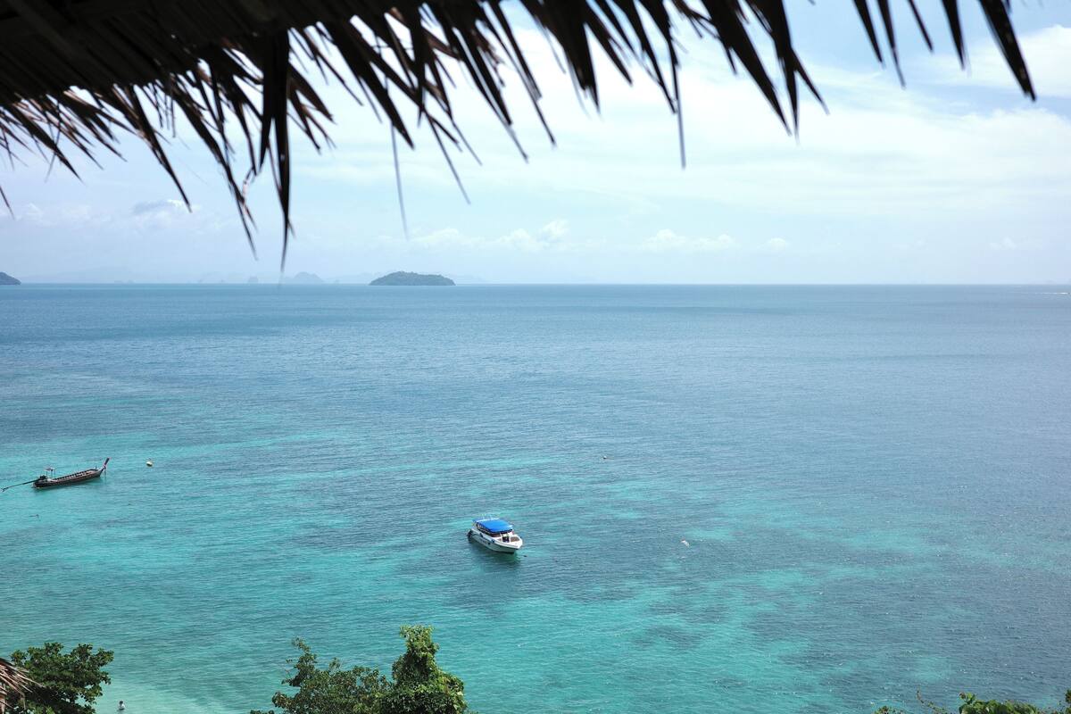 Un bateau sur une eau turquoise et limpide, avec une île lointaine et une structure au toit de chaume au premier plan.