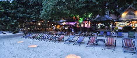 Un resort frente a la playa con sillones de descanso, un bar tiki y un edificio con techo de paja.