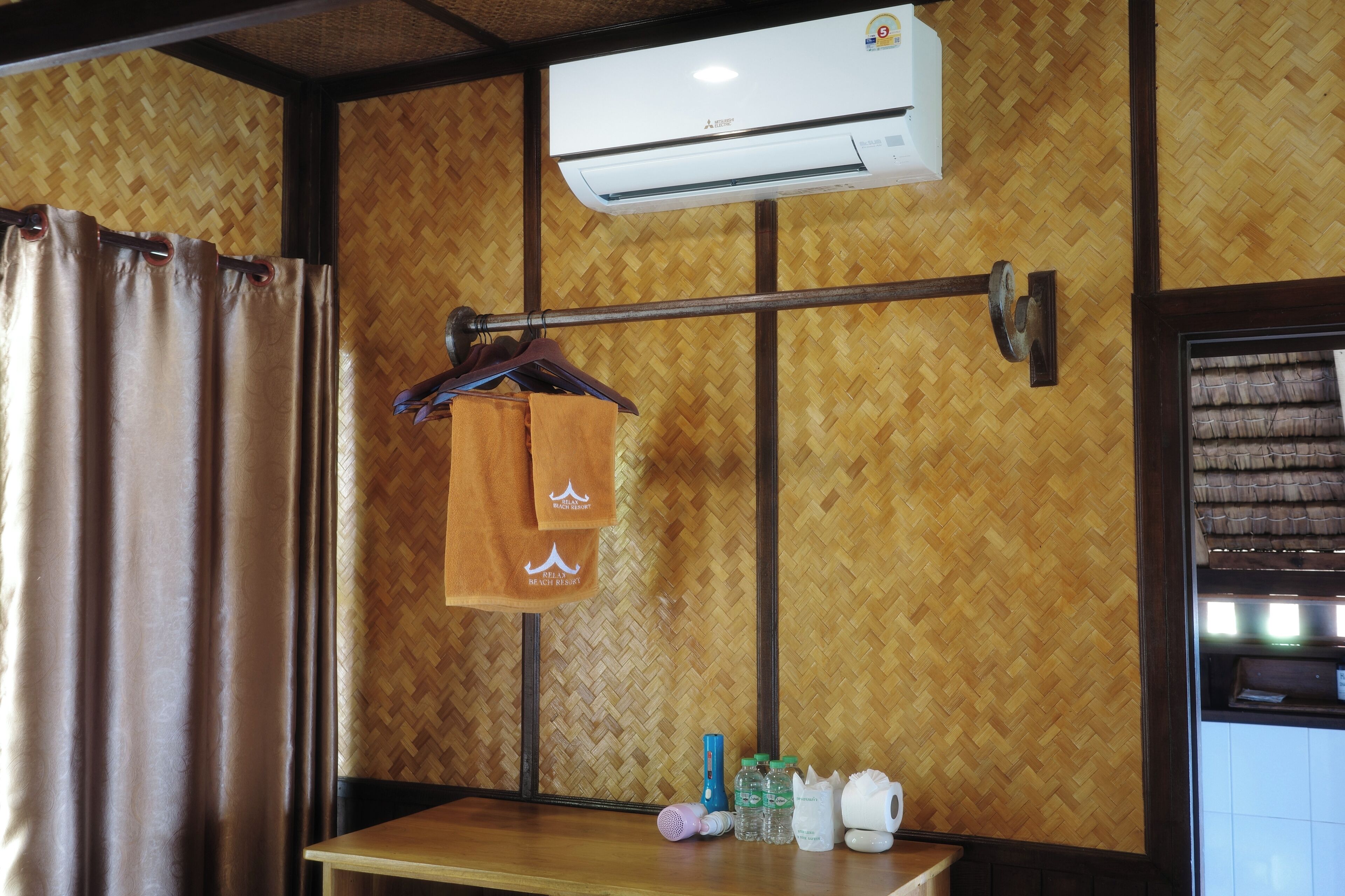 A room with a wooden wall, an air conditioning unit, a towel rack with towels, a table with toiletries, and a window with curtains.