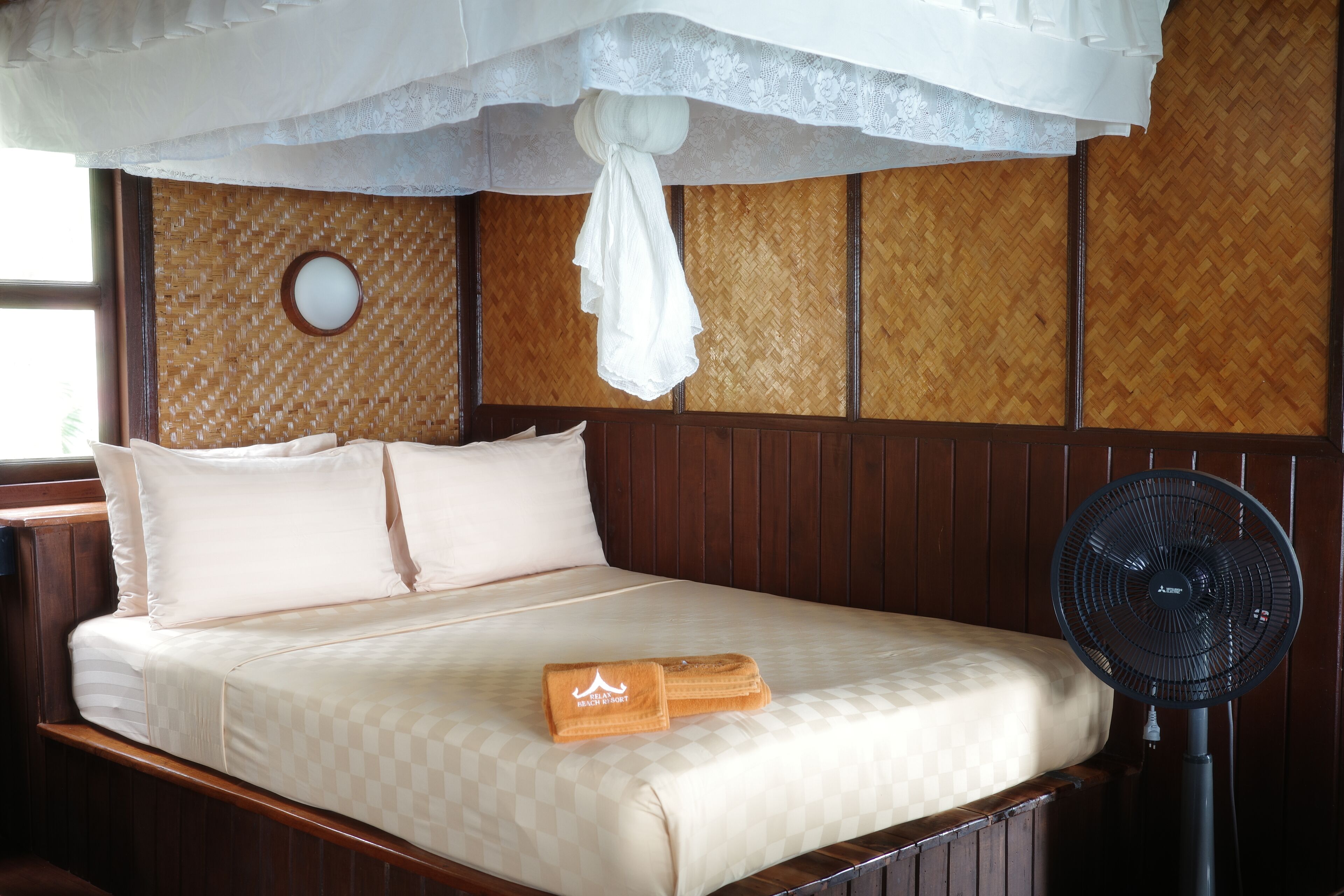 A neatly made bed with a checkered bedspread, a folded towel, and a standing fan.