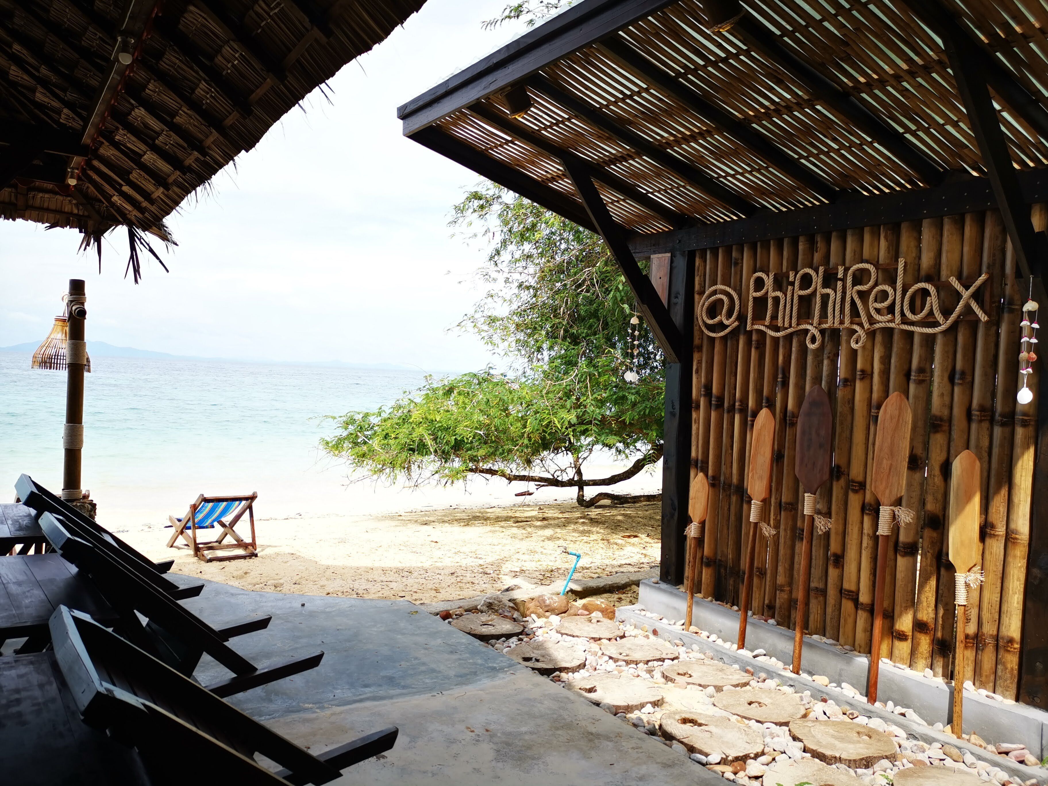 A beachside wooden structure with a thatched roof, a wooden wall with a sign, and a bench area with a view of the sea.
