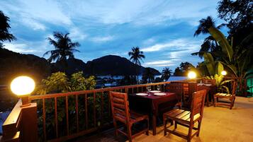 A wooden deck with tables and chairs, surrounded by tropical plants and palm trees, under a night sky.